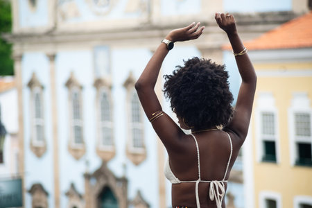 Young black woman with afro hairstyle, back to the camera, arms raised. Against old houses in the...