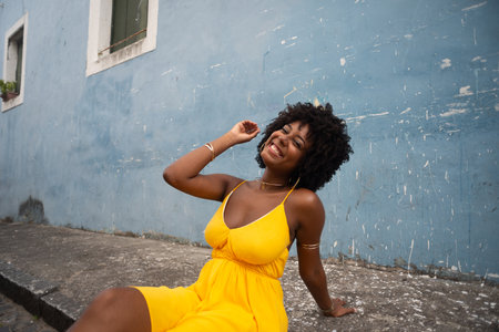 Portrait of beautiful woman wearing yellow outfit and afro hairstyle sitting on sidewalk of old street against blue wall. Confident and happy person.の写真素材