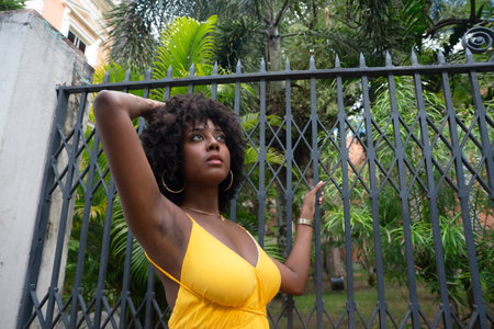 Portrait of a beautiful black woman with afro hair wearing a yellow dress leaning against an old iron fence. Happy person on a trip.の写真素材