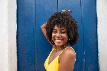 Close-up portrait of a young woman with afro hair, wearing a yellow dress, sitting by a blue door of...