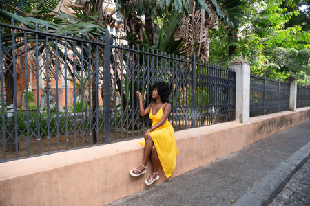 Happy smiling woman with afro hair wearing a yellow dress sitting next to an old iron fence. Happy person on a trip.の写真素材