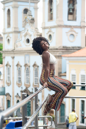 Young black woman with afro hairstyle sitting on a stainless steel railing against a church and houses in the background. Happy and empowered person. Brazilの写真素材