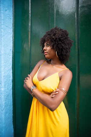 Happy smiling woman with afro hair wearing yellow dress standing with arms crossed against green door. Happy person on trip.の写真素材