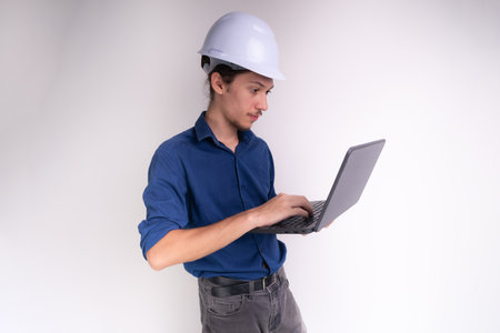 Portrait of young architect wearing white helmet, working with laptop. Isolated on white background. Safety equipment.の写真素材