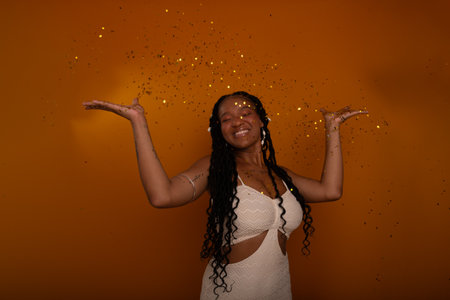 Young birthday girl wearing white outfit, happy and smiling among party confetti. Isolated against dark orange colored background. Happy and confident person.の写真素材