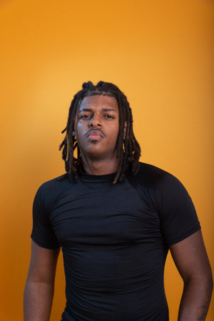 Handsome man wearing black shirt with dreadlocks posing for photo against orange background....