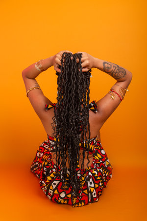 Beautiful young woman with braids in her hair, sitting with her back turned, wearing traditional clothing. Isolated on mustard colored background. Confident and calm person.の写真素材