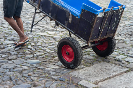 Half body of an unidentified person walking on a cobblestone street driving a handcart. Pedestrian...