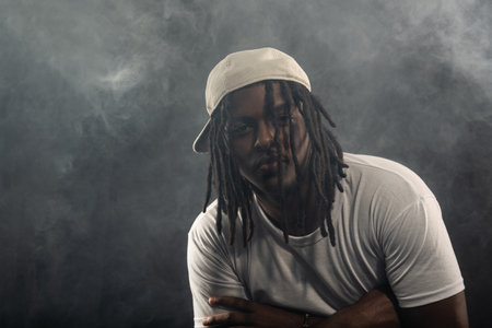 man with dreadlocks wearing a hat and white shirt posing for the camera in a studio against a smoky...