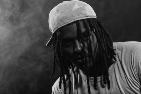 Black and white portrait of a man wearing a hat posing for the camera in a studio against a smoky...