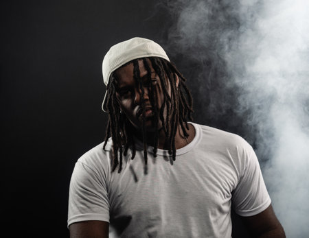 man with dreadlocks wearing a hat and white shirt posing for the camera in a studio against a smoky...
