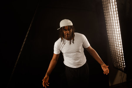 A man with dreadlocks and a white hat poses for a photo isolated on a black background in a studio.の写真素材