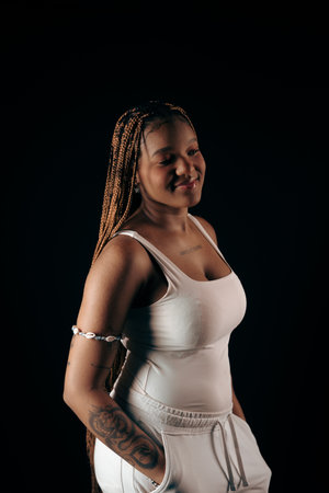 Salvador, Bahia, Brazil - August 30, 2025: Beautiful young woman with braids wearing white clothes with hands in pockets posing for the camera in studio against a black background. Salvador, Brazil.の写真素材