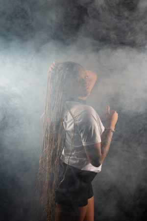 Salvador, Bahia, Brazil - August 30, 2025: Woman with braids wearing a white shirt and black shorts posing for a studio photo isolated on a smoky background. Salvador, Brazil.の写真素材