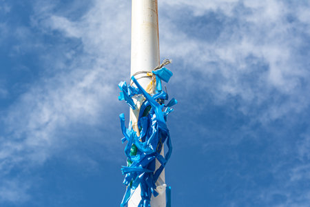 The image shows a white pole with fragments of a torn and shredded blue flag attached to it. The sky is clear, with few clouds, forming a vibrant blue background.の写真素材