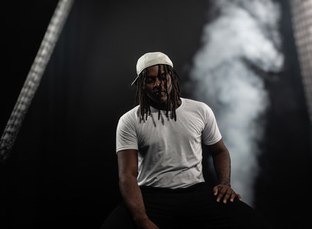 man with dreadlocks wearing a hat and white shirt posing for the camera in a studio against a smoky...