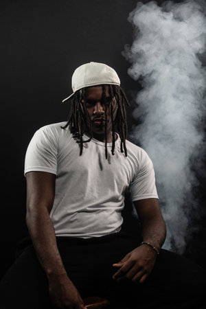 man with dreadlocks wearing a hat and white shirt posing for the camera in a studio against a smoky...