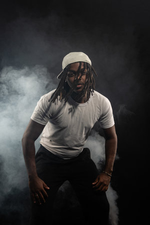 man with dreadlocks wearing a hat and white shirt posing for the camera in a studio against a smoky background. Confident and serious young man.の写真素材