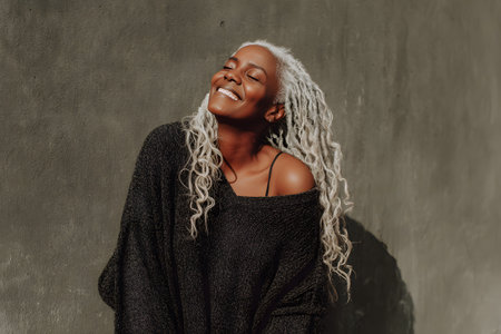 Portrait of a middle-aged or mature African American woman with a radiant smile, eyes closed, enjoying the warmth and light of the sun. The background is a simple, textured concrete wall in a dark gray tone, creating a dramatic contrast with the warm light illuminating her face and hair, enhancing happiness and confidence.の素材