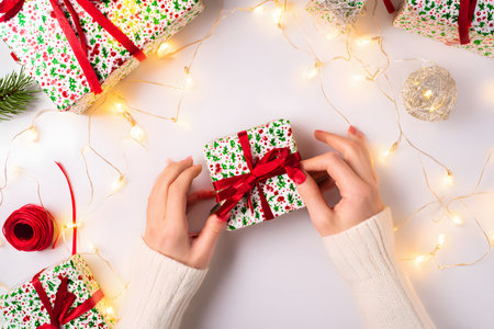 Top view of hands wearing cream sweaters tying a red ribbon around a small gift wrapped in Christmas-themed patterned paper.の素材