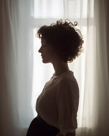 Artistic and minimalist silhouette of a pregnant woman in profile, against a light, softly gradient background. The woman holds her belly with her hands.の素材