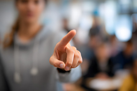 A close-up of an outstretched hand, with the index finger pointing forward, suggesting a gesture of instruction, communication, or Sign Language, with the classroom and teacher blurred in the background.の素材