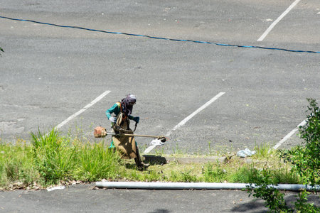 Salvador, Bahia, Brazil - November 28, 2025: A city hall employee is seen mowing tall grass in the Sao Raimundo parking lot. Salvador, Bahia.のeditorial素材