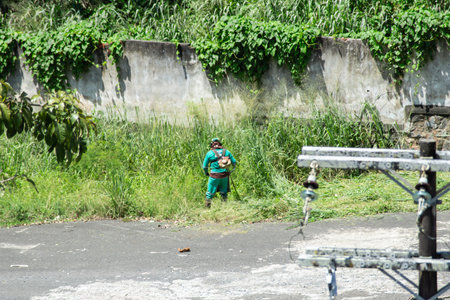 Salvador, Bahia, Brazil - November 28, 2025: Maintenance and cleaning worker in Salvador is seen mowing weeds in a parking lot. Bahia, Brazil.のeditorial素材