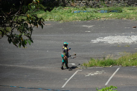 Salvador, Bahia, Brazil - November 28, 2025: A city hall employee is seen mowing tall grass in the Sao Raimundo parking lot. Salvador, Bahia.のeditorial素材