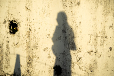Elongated silhouette of a person standing, possibly using a phone or holding something, projected onto an old, worn, light-colored wall.の写真素材
