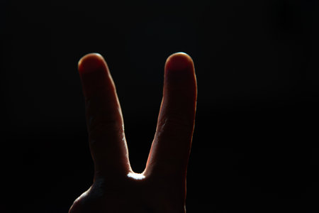 Hand in backlight making the two-finger sign, peace or victory, against a dark background. Edges of the fingers illuminated by a strong light.の写真素材