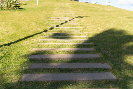 View from below of a rustic staircase made of discreet stone steps, ascending a grassy hill under the sunlight, with elongated shadows creating patterns.の写真素材