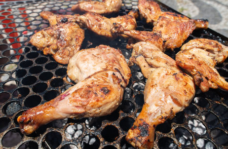 Pieces of chicken being grilled on a metal grill with circular holes in a street party setting.