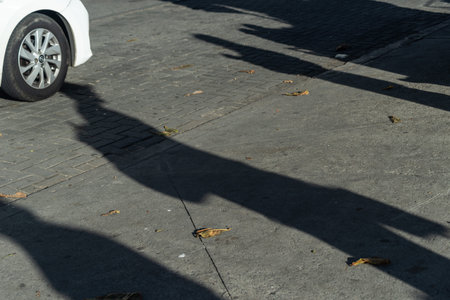 Projection of long shadows of people walking on a cobblestone sidewalk near a white car.