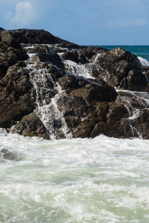 A photograph showing seawater receding and cascading like white foam down a dark, wet rocky cliff after a wave breaks.の写真素材