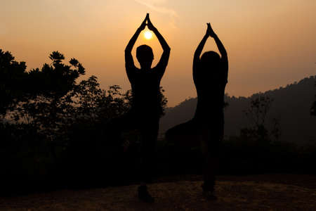 Two women doing yoga tree pose in silhouette on serene hill summit during sunriseの写真素材