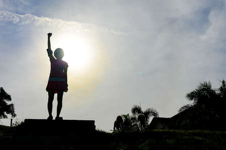 Silhouette woman with dogs hiker raises hands on hill peak against blue sky and sun, signify successの写真素材