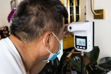 Asian man checking forehead temperature using infrared thermometer before enter establishment with focus on thermometer, as part of new normal requirementの写真素材