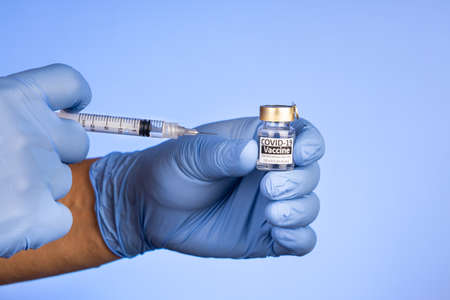 Close-up of hand with surgical glove holding covid-19 vaccine vial on blue background