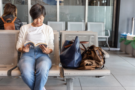 MIddle age Asian woman with luggage reading book to kill time while waiting for departure in airport terminalの写真素材