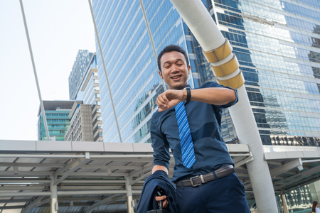 Confident young man holding suit with Briefcase and looking at his watch while standing outdoors with cityscape in the background, In time appointment Concept.の写真素材
