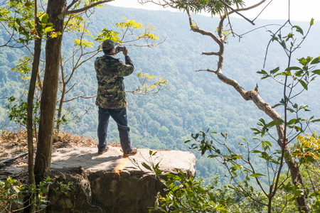 Male or Men Stand on Cliff and Take photograph of Beautiful Nature over Green Forest Mountain with Camera as Journey to Freedom Lifestyle Concept.の写真素材