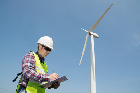 Electric Engineer writing Maintenance report on Clipboard with Wind turbine power Generator Tower Background as Green energy or Renewable Energy Technology Project Development Concept.の写真素材