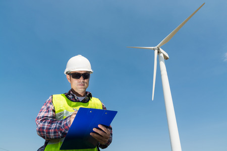 Electric Engineer writing Maintenance report on Clipboard with Wind turbine power Generator Tower Background as Green energy or Renewable Energy Technology Project Development Concept.の写真素材
