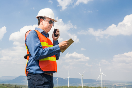 Male Architect or Engineer, use Hand-Held Transceiver Radio and Digital Wireless Tablet Device, working at Wind Turbine Power Generator Field as Infrastructure Construction Project Developmentの写真素材