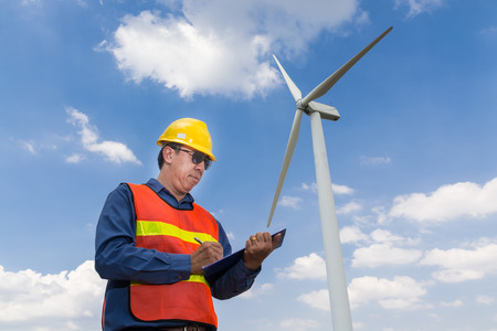 Electric Engineer writing Maintenance report on Clipboard with Wind turbine power Generator Tower Background as Green energy or Renewable Energy Technology Project Development Concept.の写真素材
