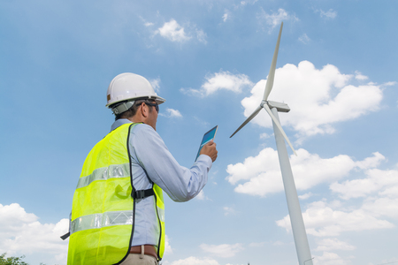 Electric Engineer writing Maintenance report on Clipboard with Wind turbine power Generator Tower Background as Green energy or Renewable Energy Technology Project Development Concept.の写真素材