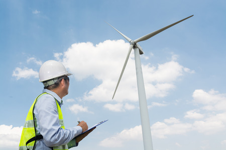 Electric Engineer writing Maintenance report on Clipboard with Wind turbine power Generator Tower Background as Green energy or Renewable Energy Technology Project Development Concept.の写真素材