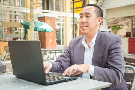 Asian Businessman typing on Notebook or Laptop Computer on Table in Outdoor Public as Mobile Workplace and Modern Lifestyleの写真素材