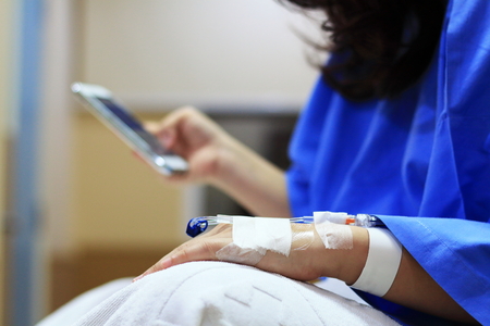 The woman patient in the hospital in blue dress in bed. Seeing the left hand was intravenous injection by dropping bottle. Using Mobile Phone.の写真素材